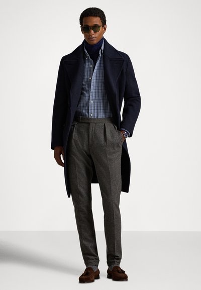 Tamsiai mėlynas vilnonis paltas, mėlyna languota palaidinė su sagomis, tamsiai pilkos klostuotos kelnės, saulės akiniai ir rudi loaferiai. Paprastas, klasikinis dizainas.