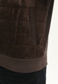 Bruine sweatshirt met een gerecycled textuur, voorzien van een ribbelzoom en zijzak. Gladde stofafwerking met een oranje accentlabel.