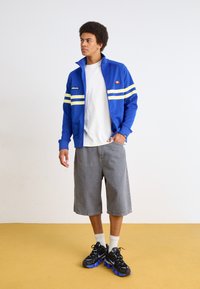 Mėlyna sportinė striukė su baltais ir geltonais dryžiais, balta marškinėliai, pilkos šortai ir juodi sportbačiai su mėlynomis akcentais. Sportbačiai turi blyksčio raštą.