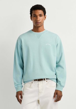 Mann trägt hellblauen Pepe Jeans Sweatshirt und weiße Hose, steht vor einem schlichten hellen Hintergrund und schaut nach vorne.