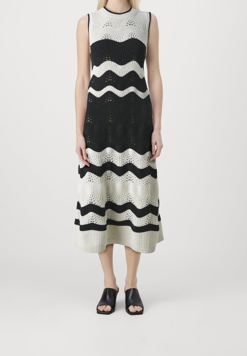 Robe midi tricotée sans manches noire et blanche avec un motif à rayures ondulées, portée avec des mules à talons ouverts noires sur un fond neutre.