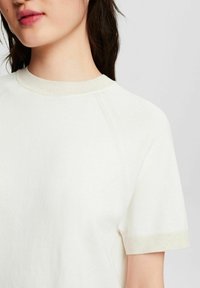 Młoda osoba z ciemnymi włosami nosi kremowy, krótki sweter z długim rękawem z prążkowanym kołnierzykiem i mankietami, skrócony na ramionach i górnej części klatki piersiowej.