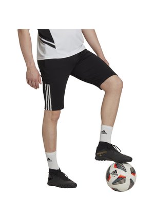 Zwarte sportshorts met drie witte strepen, gedragen met zwarte voetbal-schoenen en witte sokken, staande op een zwart-witte voetbal.