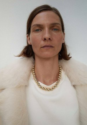 Collier de perles dorées porté sur un haut crème, associé à un manteau en fourrure légère. Le modèle a les cheveux au menton et un maquillage neutre.