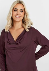 Långärmad topp i slät, djup mörkröd tyg med draperad halsringning och avslappnad passform, med subtil textur och en avslappnad stil.