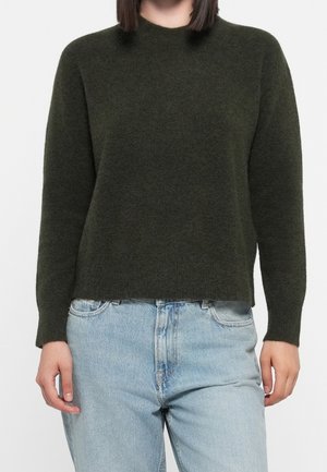 Personne portant un pull en maille à manches longues vert foncé et un jean taille haute bleu clair, debout devant un fond uni.