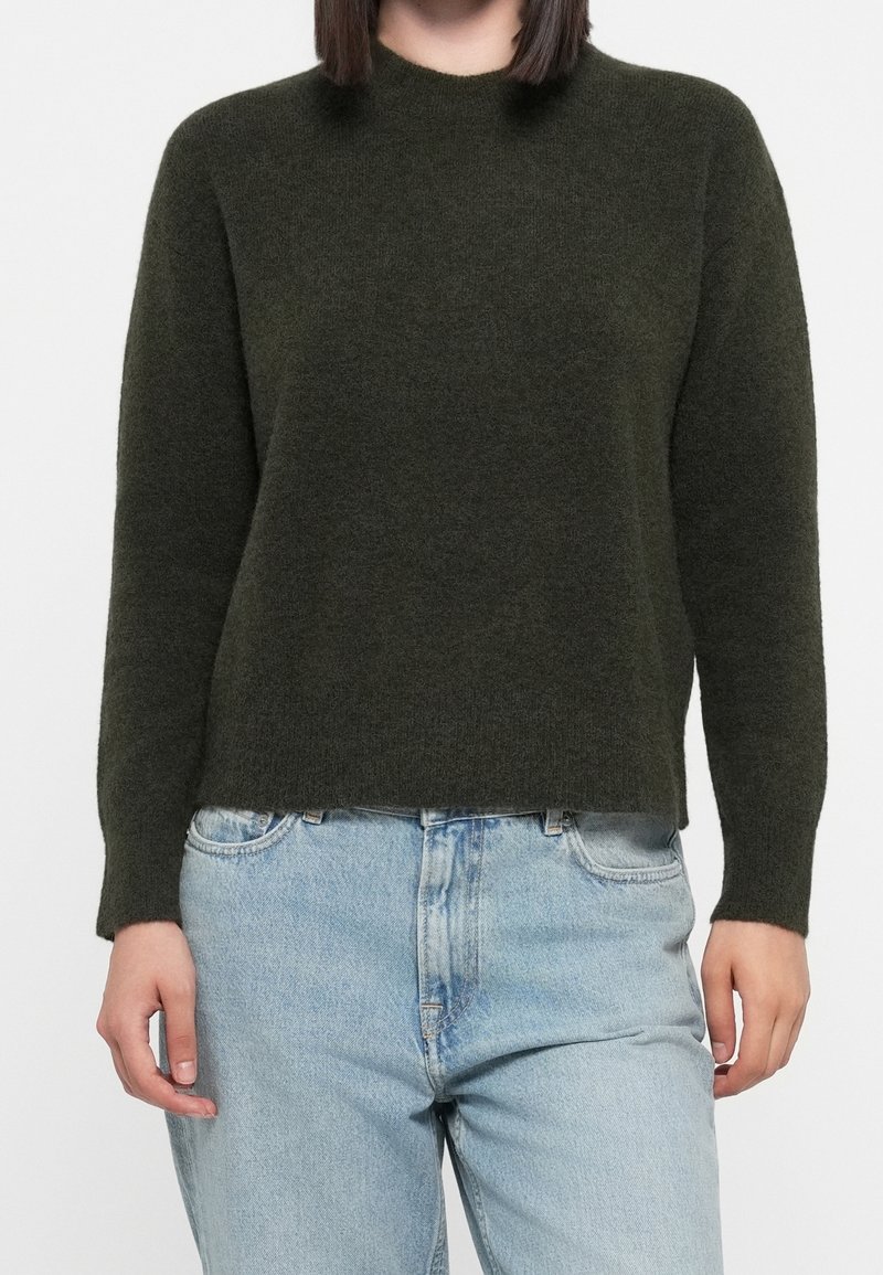 Personne portant un pull en maille à manches longues vert foncé et un jean taille haute bleu clair, debout devant un fond uni.