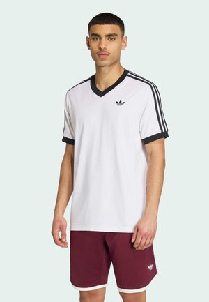 Mężczyzna ubrany w białą koszulkę Adidas z czarnymi paskami i bordowe spodenki Adidas stoi na jasnym tle