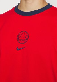 T-shirt en coton rouge avec un col côtelé bleu marine, présentant un graphisme circulaire brodé et un petit logo Nike bleu marine sur la poitrine.