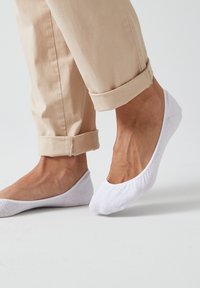 Chaussettes blanches à cheville en mélange de coton, présentant un design bas et une texture lisse. Portées avec un pantalon beige retroussé.
