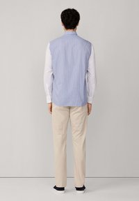 Uomo in piedi di fronte a un muro, che indossa una maglietta senza maniche a righe blu sopra una camicia a maniche lunghe bianca, pantaloni beige e scarpe da ginnastica nere.