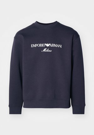 Tamsiai mėlynas megztinis su ilgomis rankovėmis, raukšlėtais rankogaliais ir apvaliu kaklu. Priekyje yra balti logotipai „EMPORIO ARMANI“ ir „Milano“.