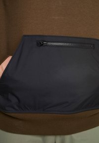 Personne portant un pull marron avec une poche avant zippée noire et la main partiellement à l'intérieur de la poche, pantalon beige visible en dessous.