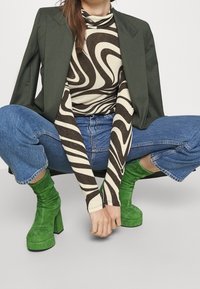 Grüne Wildleder-Stiefeletten mit hohen Blockabsätzen, kombiniert mit einem gemusterten braunen und cremefarbenen Oberteil und einem dunkelgrünen Blazer über Jeans.