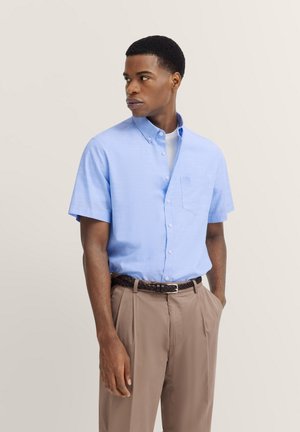 Kurzärmliges, hellblaues Hemd mit Knöpfen und Tasche, getragen über einem weißen T-Shirt, kombiniert mit hellbraunen Hosen und einem geflochtenen Gürtel.