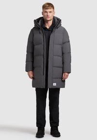 Szürke puffernadrág, közepes hosszúságú kialakítással, doboz alakú steppelt mintával, állítható kapucnival és elülső patentos záródással, sötét kapucnis pulcsi fölött viselve.