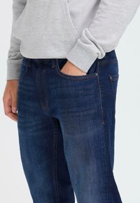 Persona vistiendo jeans azul oscuro y una sudadera gris claro, con una mano en el bolsillo de los jeans, contra un fondo liso.