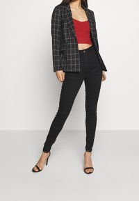 Black plaid blazer with white lines, red cropped knit top, and fitted black pants. Model wears black strappy heels and poses against a white background.