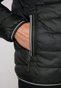 Giubbotto piumino nero con tasca laterale con zip. Le caratteristiche includono una superficie testurizzata, cuciture accentate bianche e un design aderente con polsini elasticizzati.