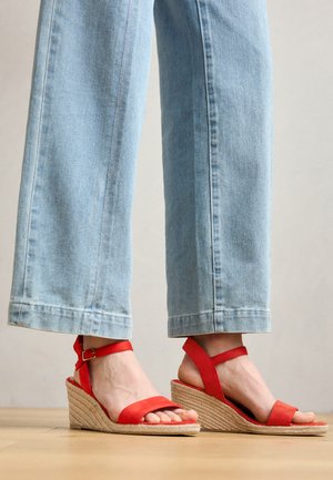 Sandales à talons en forme de coin rouges avec un talon recouvert de jute, dotées de brides autour de la cheville et de larges bandes en tissu sur les orteils. Associées à un jean cropped bleu clair.