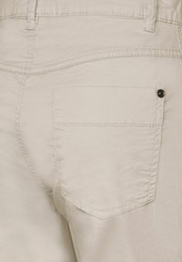 Beiga bomullbyxor med en bakficka prydd med en dekorativ knapp och subtila sömdetaljer. Tyget har en lätt lyster.