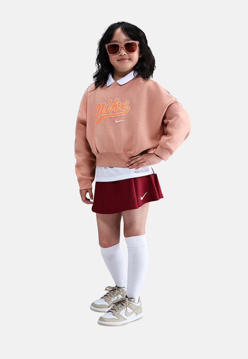 Sudadera rosa con logo de Nike naranja, camisa blanca con cuello, falda pliegues roja, calcetas blancas hasta la rodilla y zapatillas Nike beige.