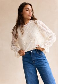Top in pizzo con ricami floreali, maniche trasparenti e scollatura arricciata, abbinato a jeans blu a vita alta. Colore beige chiaro.