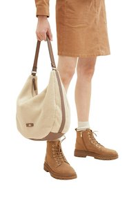 Beige nepsjaal hobo-tas met bruine leren accenten. Het model draagt tan-kleurige laarzen en een corduroy jurk, terwijl ze de tas met één hand vasthoudt.