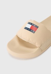 Sandal en slide beige avec une tige rembourrée, arborant un patch logo noir, rouge et blanc. Semelle lisse et mate avec des bords arrondis.