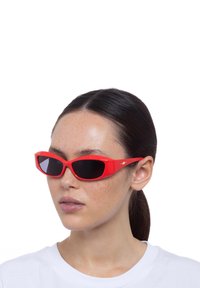 Occhiali da sole in plastica rossa con forma a occhio di gatto angolare, lenti nere e un piccolo accento del logo dorato sul lato. Modello che indossa una camicia bianca.