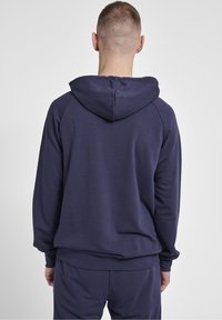 Mann mit kurzen Haaren, der einen navyblauen Hoodie und passende Hosen trägt, steht mit dem Rücken zu einem einfachen hellen Hintergrund.