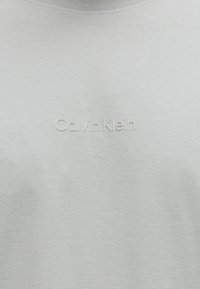 Ανοιχτό γκρι ύφασμα με ανάγλυφο λογότυπο "Calvin Klein" στο κέντρο της περιοχής του στήθους μιας απλής μπλούζας.
