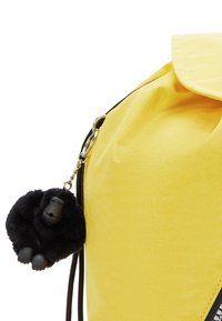 Gul ryggsäck med en svart, fluffig gorillanyckelring fäst nära den övre dragkedjan.