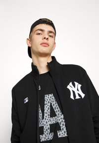 Sort zip-up jakke med New York Yankees logo. Båret over en sort skjorte med et gråt plet-farvet 'LA' grafik. Blødt stof, afslappet design.