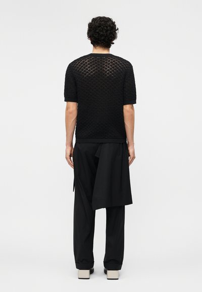 Άνδρας με σγουρά μαλλιά, φορώντας μαύρο πλεκτό τοπ με κοντά μανίκια και φαρδιά μαύρη παντελόνα με ασύμμετρη λεπτομέρεια υφάσματος στη δεξιά πλευρά.