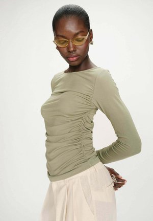 Femme portant un haut vert clair froncé à manches longues et un pantalon plissé crème, portant des lunettes jaunes œil de chat et des boucles d'oreilles en or, posant devant un fond blanc.