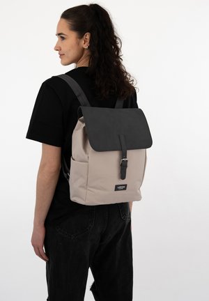 Beige backpack with a black flap and buckle closure, featuring grey straps. Material appears smooth with a structured design.