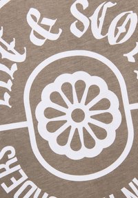 Λευκό ανθικό έμβλημα με στρογγυλεμένα πέταλα μέσα σε οβάλ, περιτριγυρισμένο από μερικό λευκό κείμενο σε υφασμένο μπεζ φόντο.