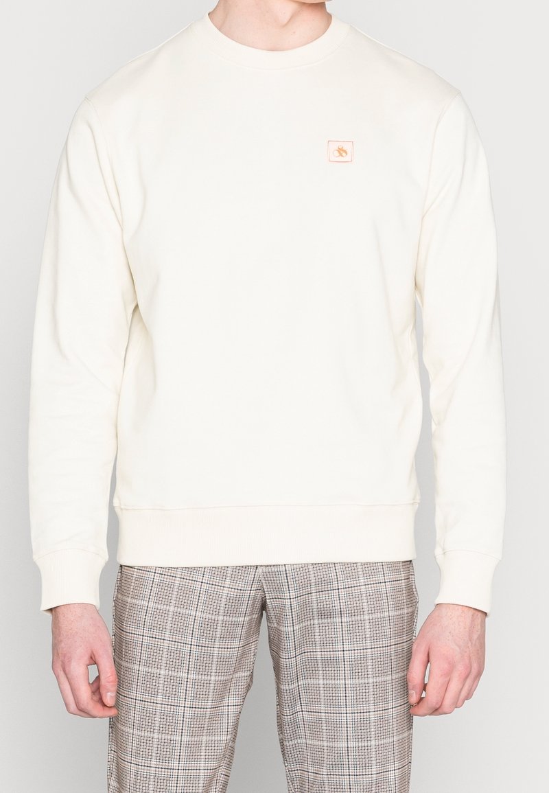 Sudadera de color crema hecha de tela suave, con un cuello redondo y puños acanalados, y un pequeño logotipo bordado en el pecho.
