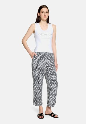 Young woman standing wearing white sleeveless top with "GROW UP" text, black and white geometric pattern pants, and black slide sandals.