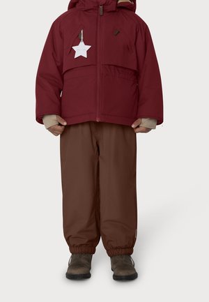 Enfant portant une veste d'hiver à capuche bordeaux avec une étiquette blanche en forme d'étoile, pantalon marron, manches intérieures beiges et bottes marron.