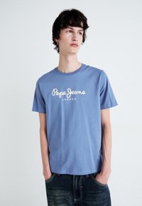 Jaunas vyras su garbanotais tamsiais plaukais, vilkintis mėlyną "Pepe Jeans London" marškinėlį ir tamsias džinsas, stovi su rankomis kišenėse.