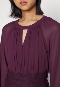 Femme portant une robe prune à manches longues avec un décolleté en trou de serrure et un détail en dentelle à la taille, visible des épaules jusqu'au milieu du torse.