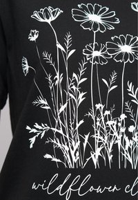 T-shirt noir avec un dessin en ligne blanche de fleurs sauvages et d'herbe, avec le texte "wildflower" en police cursive en bas.