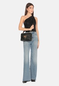 Bolso de mano negro acolchado con herrajes dorados, que presenta un cierre prominente, combinado con una blusa negra de un solo hombro y jeans de pierna ancha de color azul claro.