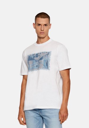 Camiseta de algodón blanca con un diseño gráfico azul que incluye el texto "GUESS" y "LOS ÁNGELES", cuello redondo y mangas cortas.