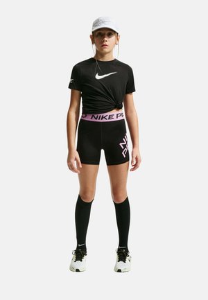 Joven atleta femenina que lleva una camiseta negra anudada de Nike, pantalones cortos Nike Pro negros y rosas, calcetines negros hasta la rodilla, zapatillas blancas y una gorra blanca.