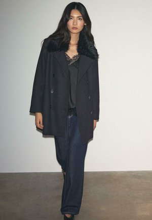 Sort dobbeltradet frakke med falsk pelskrave, båret over en sort lace-top og mørke denimbukser. Glat tekstur, afslappet silhuet.