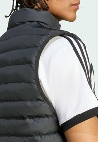 Person iført en sort quiltet vest over en hvid t-shirt med tre sorte striber på skulderen, vendt sideways.