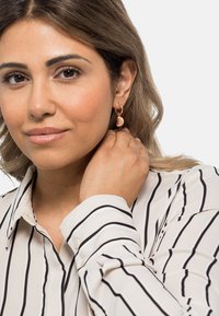 Orecchini a cerchio in oro rosa con un design curvato e semisferico, pendenti dal lobo dell'orecchio, indossati con una camicia bianca a righe nere.